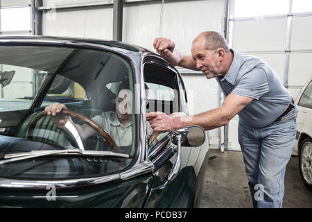 Un blanc senior male mécanicien de voiture montre son petit-fils de la vue du conducteur Le siège d'une vieille voiture dans une voiture classique atelier de réparation. Banque D'Images