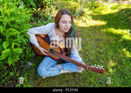 Jeune femme assise hipster dans l'herbe et jouer de la guitare sur parc ou jardin arrière-plan. Teen girl apprendre à jouer la chanson et de la musique. Passe-temps, de vie, de vous détendre, d'un instrument, l'éducation, loisirs concept Banque D'Images