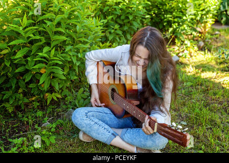 Jeune femme assise hipster dans l'herbe et jouer de la guitare sur parc ou jardin arrière-plan. Teen girl apprendre à jouer la chanson et de la musique. Passe-temps, de vie, de vous détendre, d'un instrument, l'éducation, loisirs concept Banque D'Images