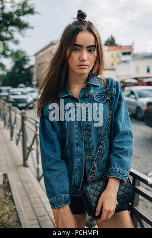 Jeune femme aux cheveux longs habillé en veste en jean debout sur la rue près de la route. Cute teen girl posing élégant dans la ville. Banque D'Images