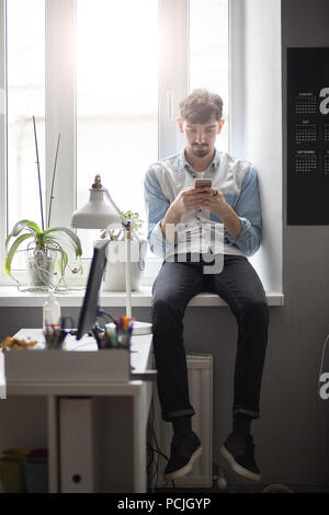 Élégant jeune homme assis sur la fenêtre avec le téléphone dans les mains Banque D'Images