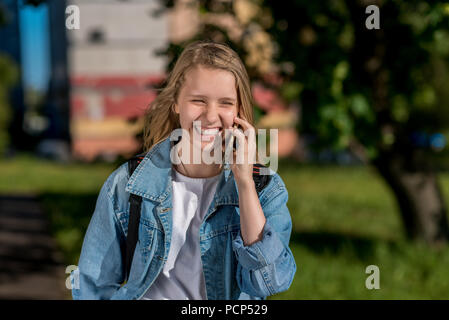 Jeune fille lycéenne à parler au téléphone, en été dans la nature, souriant joyeusement, émotionnellement heureux. Dans ses mains est titulaire d'un smartphone, en veste en jean. Banque D'Images