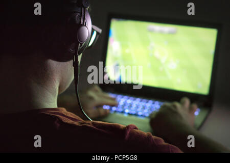 Jouer à des jeux vidéo avec l'ordinateur portable. Jeune homme joue en ligne soccer ou de football sur ordinateur. Vue arrière de gamer avec des écouteurs dans le noir ou tard dans la nuit. Banque D'Images