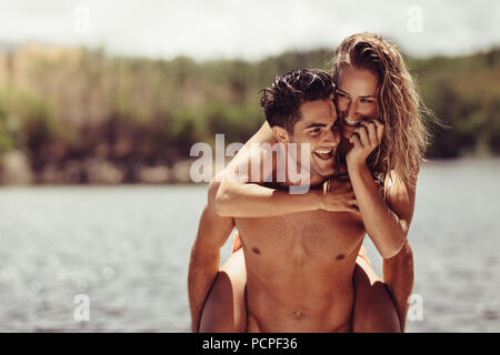 Beau jeune homme portant sa belle petite amie sur le dos par le lac. Couple leurs vacances à l'extérieur. Piggyback ride fun. Banque D'Images