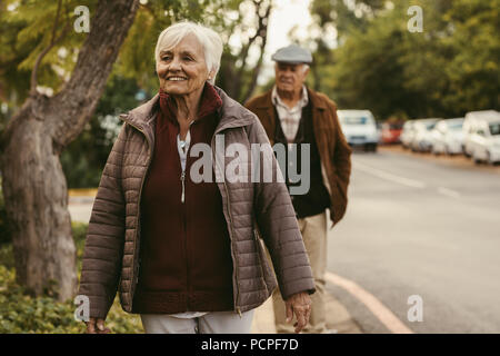 Woman veste chaude la marche à l'extérieur avec son mari à l'arrière sur journée d'hiver. Heureux femme âgée à pied par la route. Banque D'Images