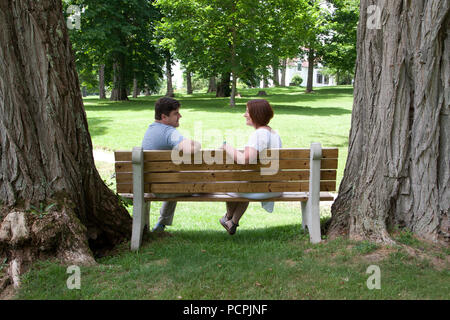 Un homme et une femme s'asseoir ensemble sur un banc de parc entouré d'arbres immenses, à la recherche à l'un l'autre et souriant Banque D'Images