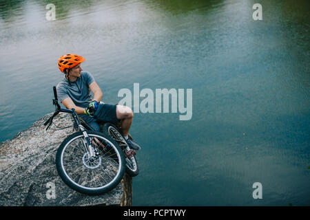 Beau jeune motard de première détente sur une falaise rocheuse au-dessus de l'eau Banque D'Images