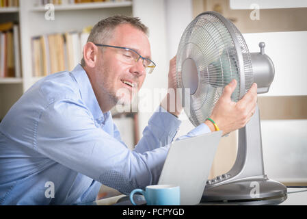 L'homme souffre de la chaleur tout en travaillant dans le bureau et tente de se rafraîchir par le ventilateur Banque D'Images