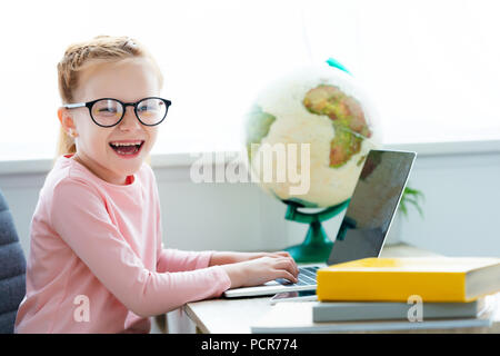 Adorable écolier heureux en lunettes étudie avec ordinateur portable et des livres à la maison Banque D'Images