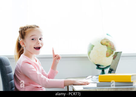 Cheerful redhead enfant pointe vers le haut avec le doigt et smiling at camera tout en étudiant avec des livres et l'ordinateur portable à la maison Banque D'Images