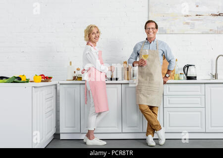 Happy senior couple smiling at camera tabliers dans tandis que boire du vin et de la cuisson dans la cuisine Banque D'Images