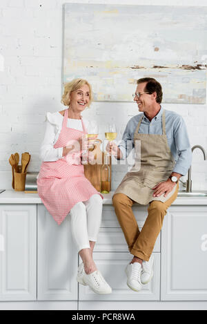 Happy senior couple holding tabliers en verres de vin tout en restant assis ensemble dans la cuisine Banque D'Images