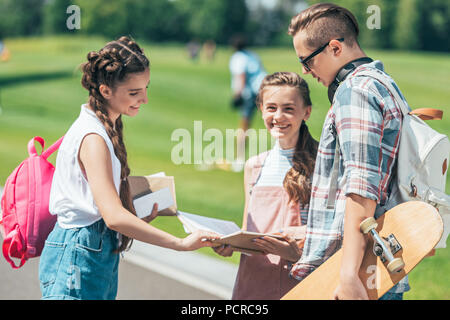 Smiling teenage students avec sacs à dos et du skateboard et permanent reading book together in park Banque D'Images