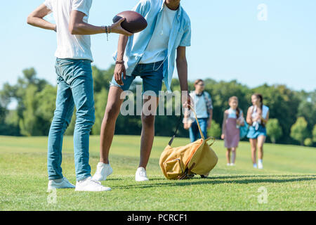 Cropped shot of multiethnic garçons jouant avec ballon de rugby pendant que leurs camarades de marcher derrière in park Banque D'Images