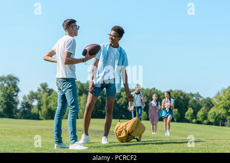 Les garçons multiethnique jouant avec ballon de rugby pendant que leurs camarades de marcher derrière in park Banque D'Images