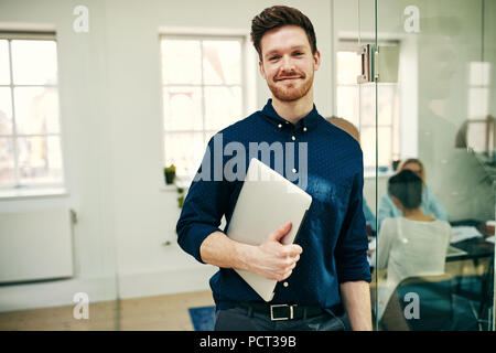 Young businessman smiling avec confiance tout en se tenant dans un bureau moderne tenant son ordinateur portable avec des collègues de travail à l'arrière-plan Banque D'Images