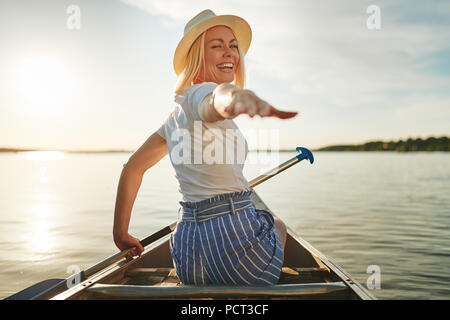 Laughing young woman looking back par dessus son épaule et s'adressant avec sa main tout en pagayant un canoë sur un lac en été Banque D'Images