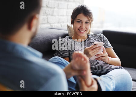 Young hispanic couple portant sur la table à la maison, à l'aide d'un téléphone mobile pour l'Internet et les médias sociaux. Le petit ami est de donner un massage de pieds de la wo Banque D'Images