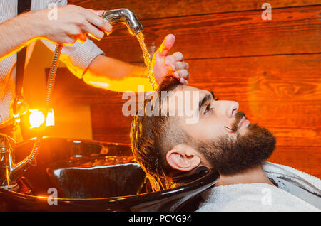 Coiffure lave la tête d'un jeune beau gars avec une barbe et moustache dans l'évier. Salon de beauté masculine. Banque D'Images