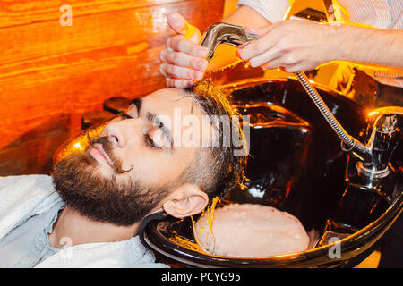 Coiffure lave la tête d'un jeune beau gars avec une barbe et moustache dans l'évier. Salon de beauté masculine. Banque D'Images