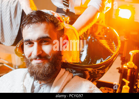 Coiffure lave la tête d'un jeune beau gars avec une barbe et moustache dans l'évier. Salon de beauté masculine. Banque D'Images