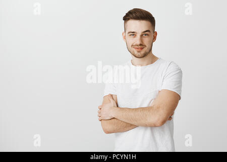 Jeune homme intrigué smart entrepreneur dans t-shirt blanc avec moustache souriant tenant les mains croisées sur la poitrine sentiment satisfait et heureux de recevoir g Banque D'Images