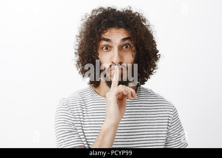 Portrait d'intigued curly attrayant brun avec barbe, excité permanent sur fond gris, disant chut chut tout en montrant l'index avec des gestes Banque D'Images