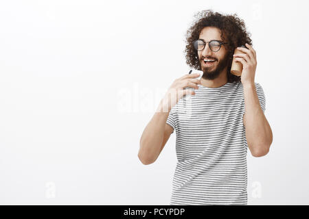 Portrait de carefree gunny bel homme avec barbe en lunettes noires, holding smartphone comme micro et chanter avec tasse de café près de l'oreille, Banque D'Images