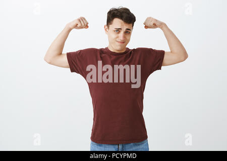 Regardez comment je suis forte. Portrait de l'auto-assurance handsome guy européenne en rouge t-shirt, soulevant les bras et montrant les muscles avec l'expression de confiance, de Bragg Banque D'Images