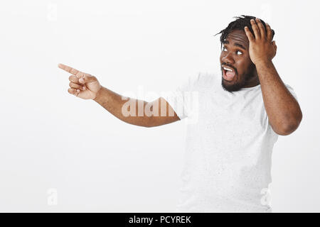 Portrait de choqué impressionné de peau foncée type en T-shirt blanc, qui s'étend à la main, et pointant vers la gauche tout en maintenant l'entendre sur palm, d'être surpris Banque D'Images