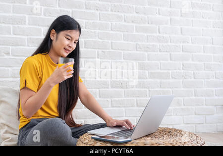 Femme d'origine asiatique s'asseoir sur le sol à l'aide de l'ordinateur portable sur support en osier et verre de jus d'orange dans la salle de séjour à la maison avec vous détendre émotion.Travailler à la maison travail concept.de Banque D'Images