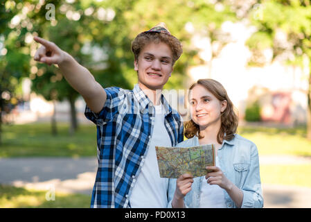 Le gars avec la fille sourit joyeusement. En été dans le parc de la nature. Dans ses mains est titulaire d'une carte de route. Avec un geste de la main et montre la direction de mouvement. Les jeunes touristes en vacances. Banque D'Images