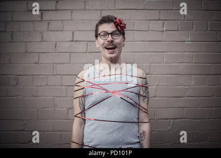 Jeune homme blanc avec des tatouages enveloppé dans un ruban cadeau rouge, avec un arc rouge dans le côté de la tête, la moustache, et lunettes vintage. Banque D'Images
