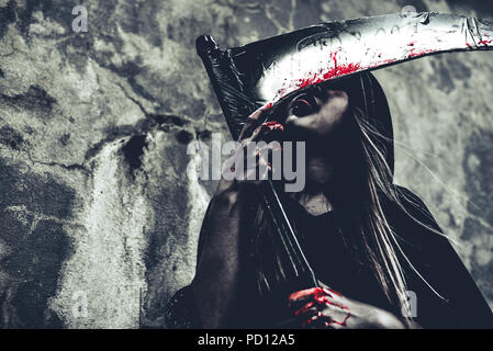 Lécher le sang sur reaper sorcière. Ange démon femelle de noir et le capot sur grunge wall background. Journée de l'Halloween et le mystère concept. Fantasme de m Banque D'Images