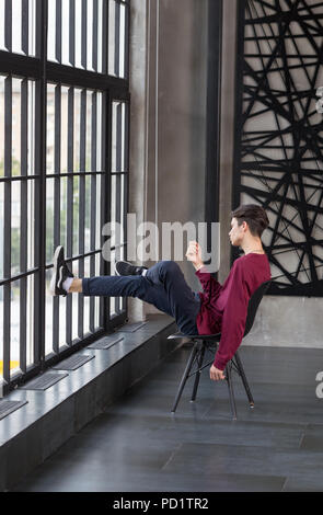 Slim beau jeune homme assis sur une chaise, pieds au repos contre la fenêtre dans un appartement branché Banque D'Images
