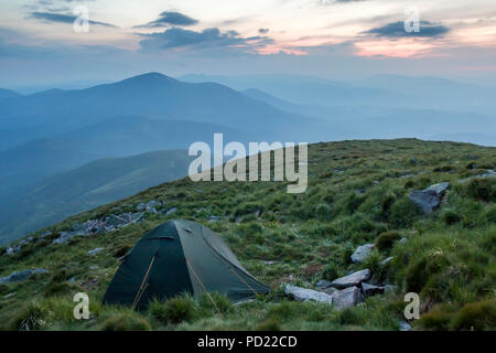 Camping d'été dans les montagnes à l'aube. Tente ronde touristique sur colline herbeuse sur bleu lointain de montagnes sous le ciel rose avant le lever ou coucher du soleil. Banque D'Images