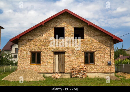 Nouveau pas fini de maison en brique avec entrée de porte en plastique à bord, fenêtres au premier étage, des tas de briques et de gravier sur l'herbe verte sur terrain clôturé. Banque D'Images