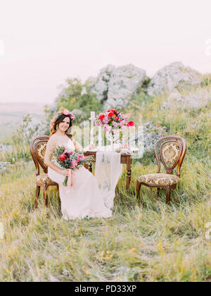 La belle mariée avec le bouwuet est assis sur la chaise ancienne près de la table de mariage dans les montagnes. Banque D'Images