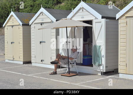 Homme assis dans une chaise lisant à l'extérieur d'une cabane de plage. Bournemouth, Dorset, Royaume-Uni, lundi 6th août 2018. Le temps chaud sur la plage de la côte sud comme la glorieuse vague de chaleur d'été continue. Banque D'Images