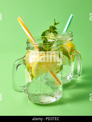 Deux bocaux Mason avec un cocktail à la main naturelle froide avec bulles d'air. Boisson rafraîchissante ingrédient naturel - agrumes, glace, feuille de menthe, de l'eau sur un fond vert. Banque D'Images