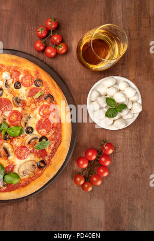 Une photo prise à la verticale d'une pizza au pepperoni, avec un verre de vin blanc, tomates cerises et mozzarellini fromage, tourné à partir de ci-dessus sur une sombre backgroun rustique Banque D'Images