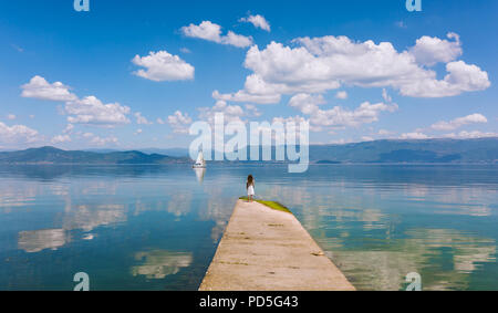 Vue panoramique sur le lac avec la réflexion de beaux nuages, une voile de navire en distance et une jeune fille regardant la scène. Banque D'Images