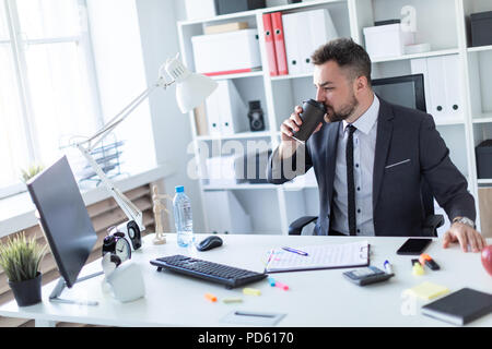 Un homme est assis dans le bureau à la table, boit du café et regarde l'écran. Banque D'Images