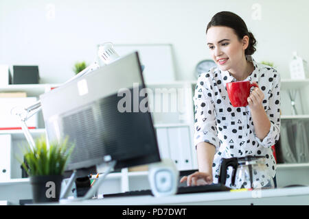 Une jeune fille se tient dans le bureau près de la table, tenant une tasse rouge dans sa main et en tapant sur le clavier. Banque D'Images