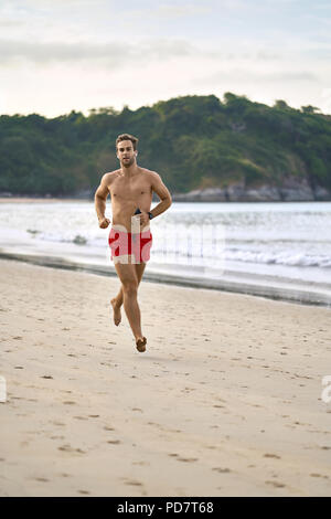 L'homme sportif bronzé est exécuté sur la plage de sable sur le fond de la mer et les collines vertes et l'ciel nuageux. Il porte un slip de bain rouge, whit Banque D'Images