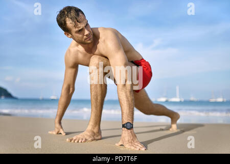 Cute homme bronzé sur la plage de sable sur le fond ensoleillé de la mer avec des bateaux blancs et le ciel bleu. Il porte un slip de bain rouge foncé et d'une montre. H Banque D'Images