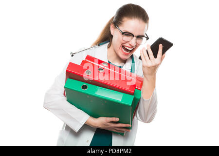 Jolie brunette caucasien angry female doctor standing in office avec des liants dans les mains sur fond blanc hurlant dans son téléphone Banque D'Images
