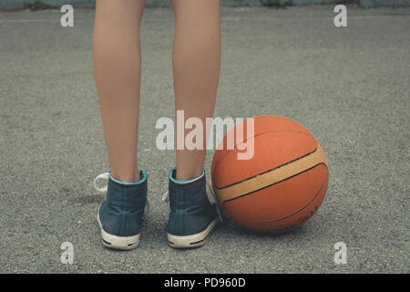 Jeune fille jambes portant des sneakers debout à côté de la balle sur le terrain de basket-ball avec une surface en béton. Low angle vue en gros plan. Banque D'Images