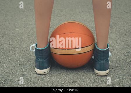 Jeune fille jambes portant des chaussures de vous tenir directement au-dessus de la balle sur un terrain de basket-ball avec une surface en béton. Low angle vue en gros plan. Banque D'Images