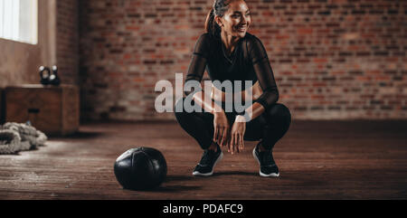 Femme souriante accroupie au studio de fitness avec un medecine ball sur le sol. femme de fitness prenant pause après l'entraînement au gymnase. Banque D'Images